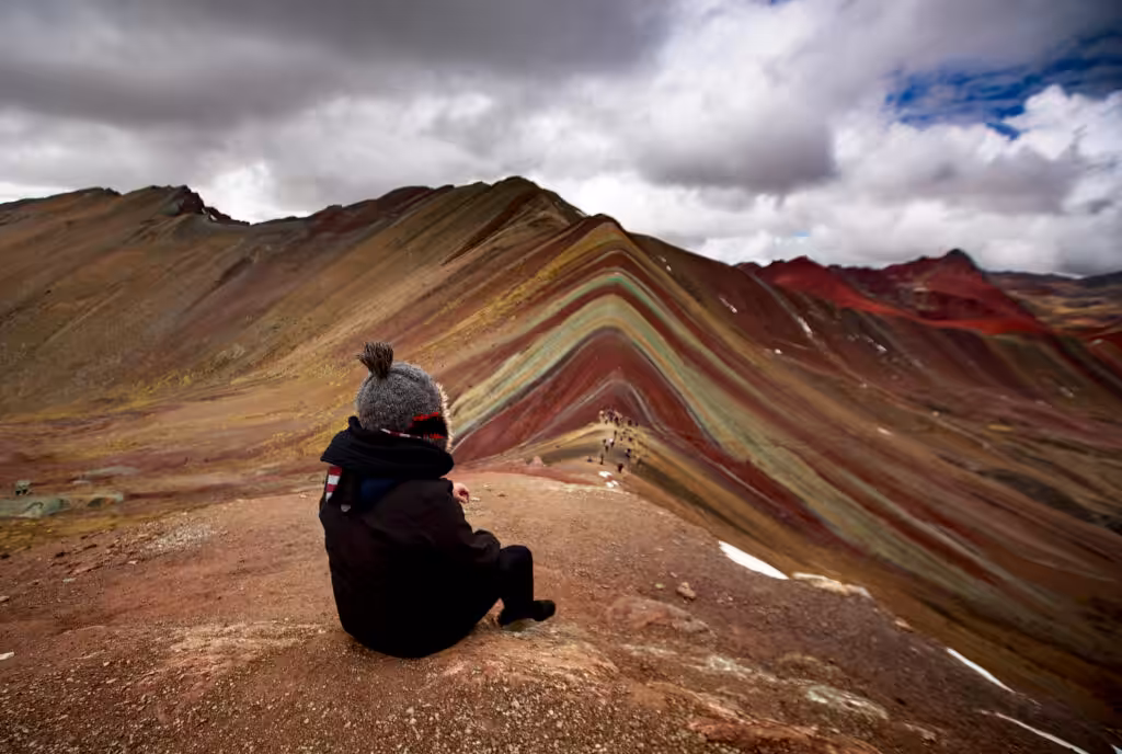 Visitante sentado no mirante com vista para a Rainbow Mountain sob céu nublado