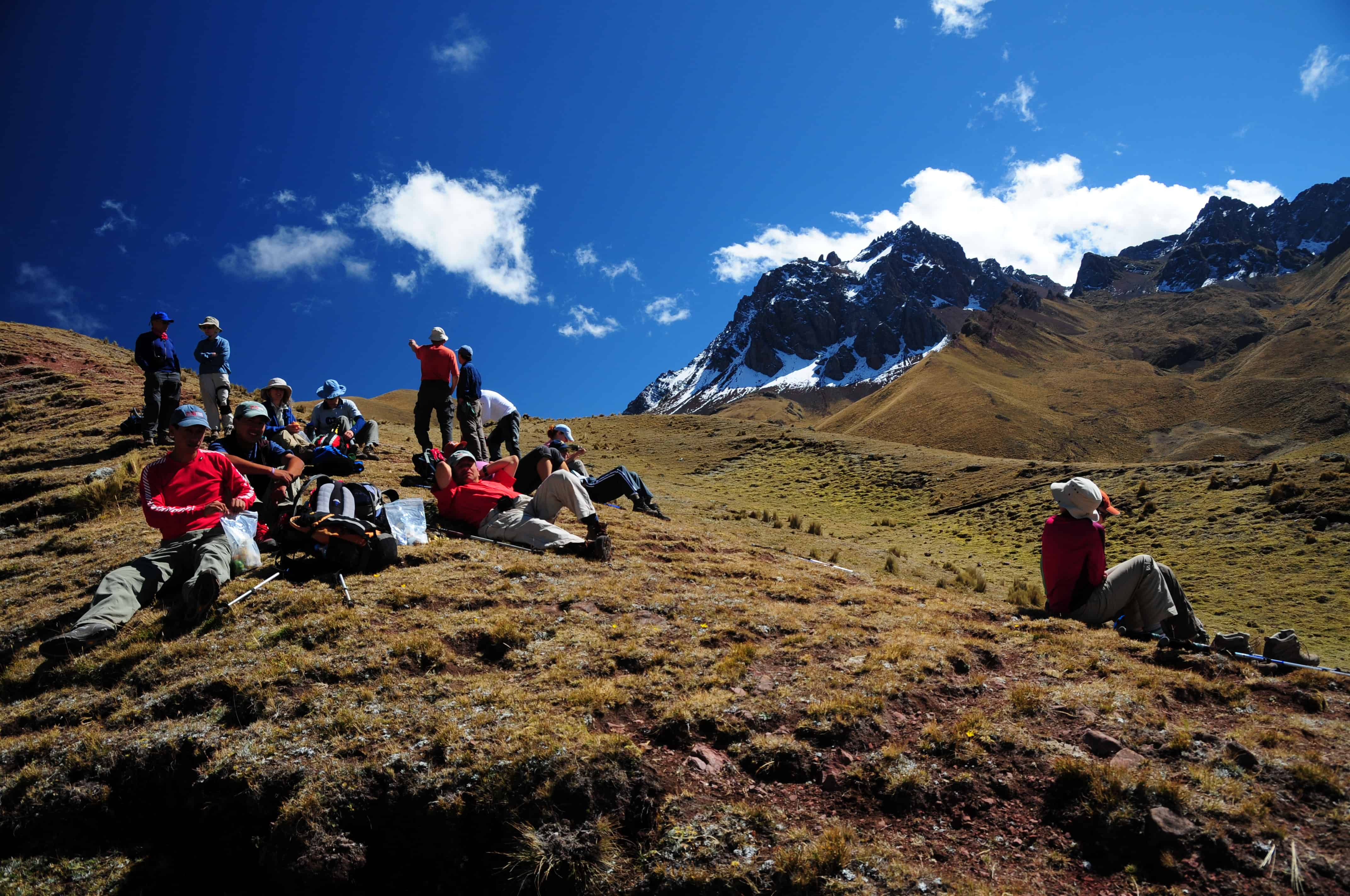 Hikers resting on grassy hill, snowy mountains background.